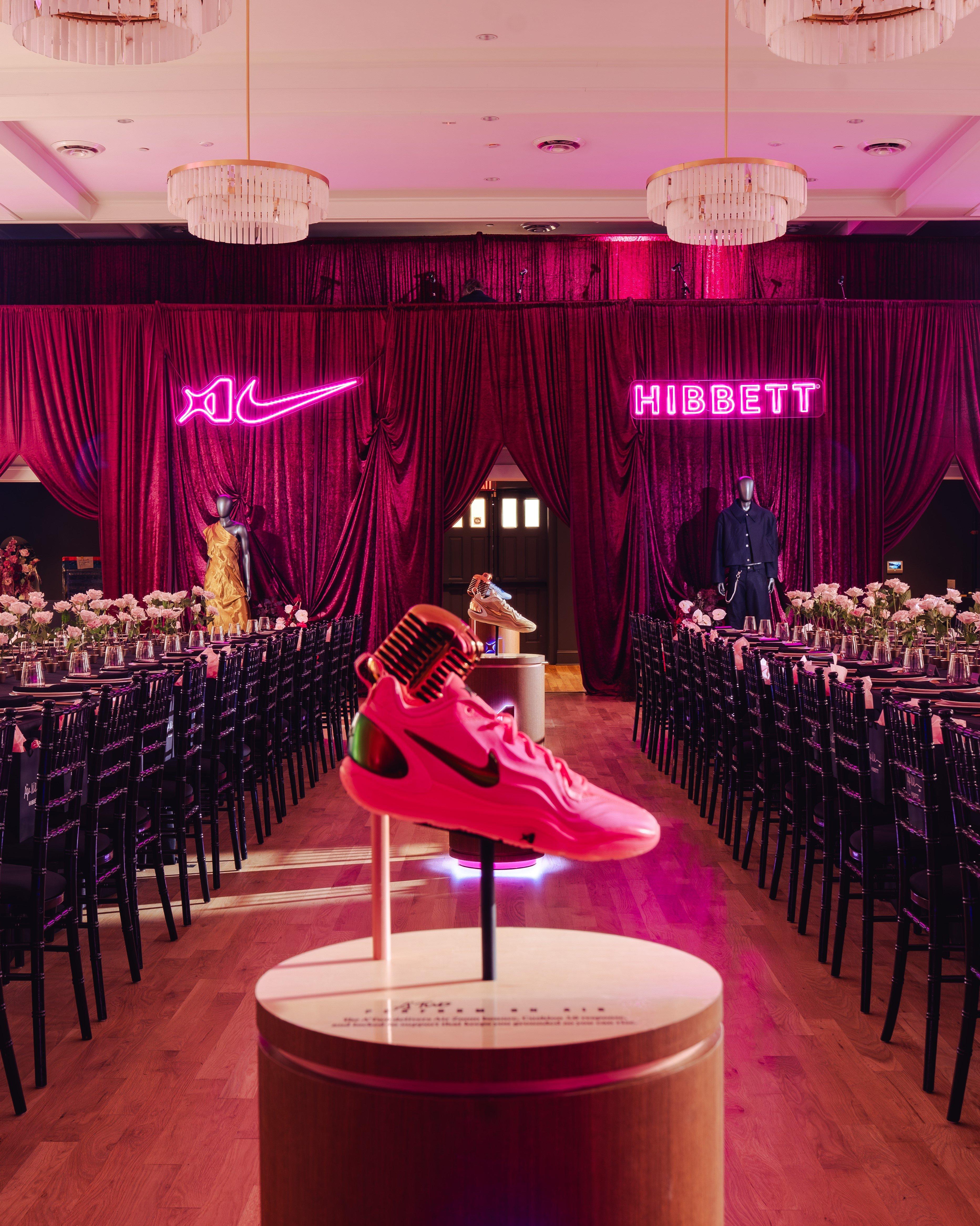 A bright pink Nike A'ja Wilson signature sneaker is displayed on a circular wooden pedestal in the center of a formal banquet room. Long dinner tables with black chairs flank the room, leading toward a stage backdrop of deep red curtains illuminated by glowing pink neon signs of the Nike Swoosh and the Hibbett logo.