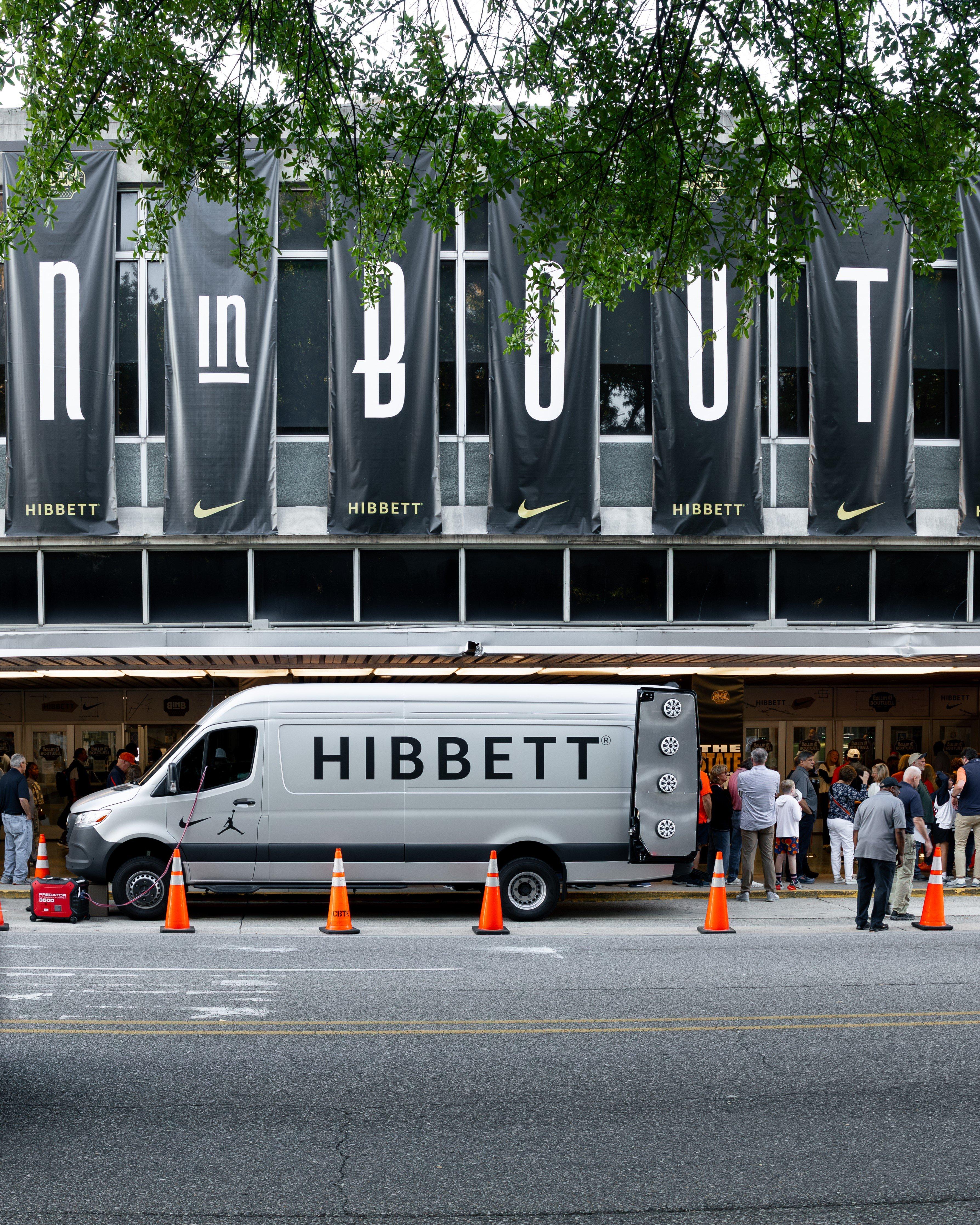 An exterior shot of an arena building with large banners hanging down, spelling out "BALLIN IN BOUTWELL." A silver HIBBETT van is parked in front, with orange traffic cones lining the street.
