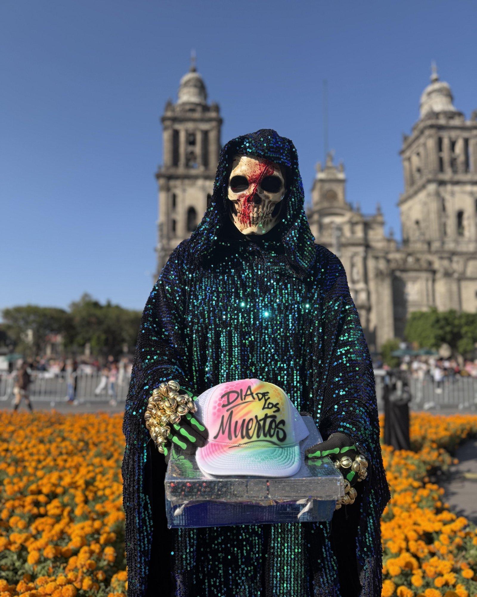 Person in a sequined skeleton costume holding a custom D&iacute;a de los Muertos airbrushed hat in front of marigold flowers and a historic cathedral.