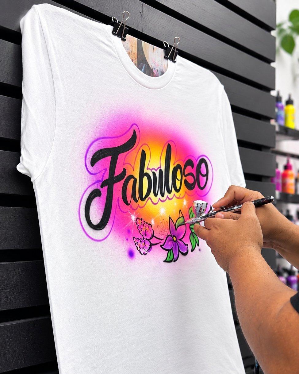 Artist airbrushing a custom &ldquo;Fabuloso&rdquo; design on a white T-shirt.