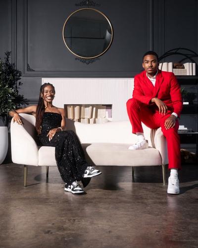 A stylish couple poses on a white couch. The woman on the left wears a strapless, sparkling black dress with her hair in long braids. The man on the right wears a bright red suit with white sneakers. They are sitting in a room with dark gray walls and a decorative fireplace.