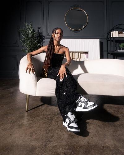 A young woman with long braids and a strapless, sparkling black outfit sits on a white couch. She wears white and black sneakers, and her hands are decorated with gold bracelets and rings. The background is a dark gray wall with white decorative molding and a round gold-framed mirror.