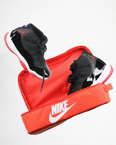 Black Air Jordan 11 sneakers with red outsoles displayed inside an open red Nike shoe storage bag.