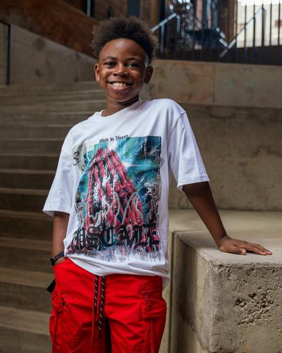 Child model wearing a white Hustle graphic T-shirt and red cargo pants while standing beside a concrete staircase.