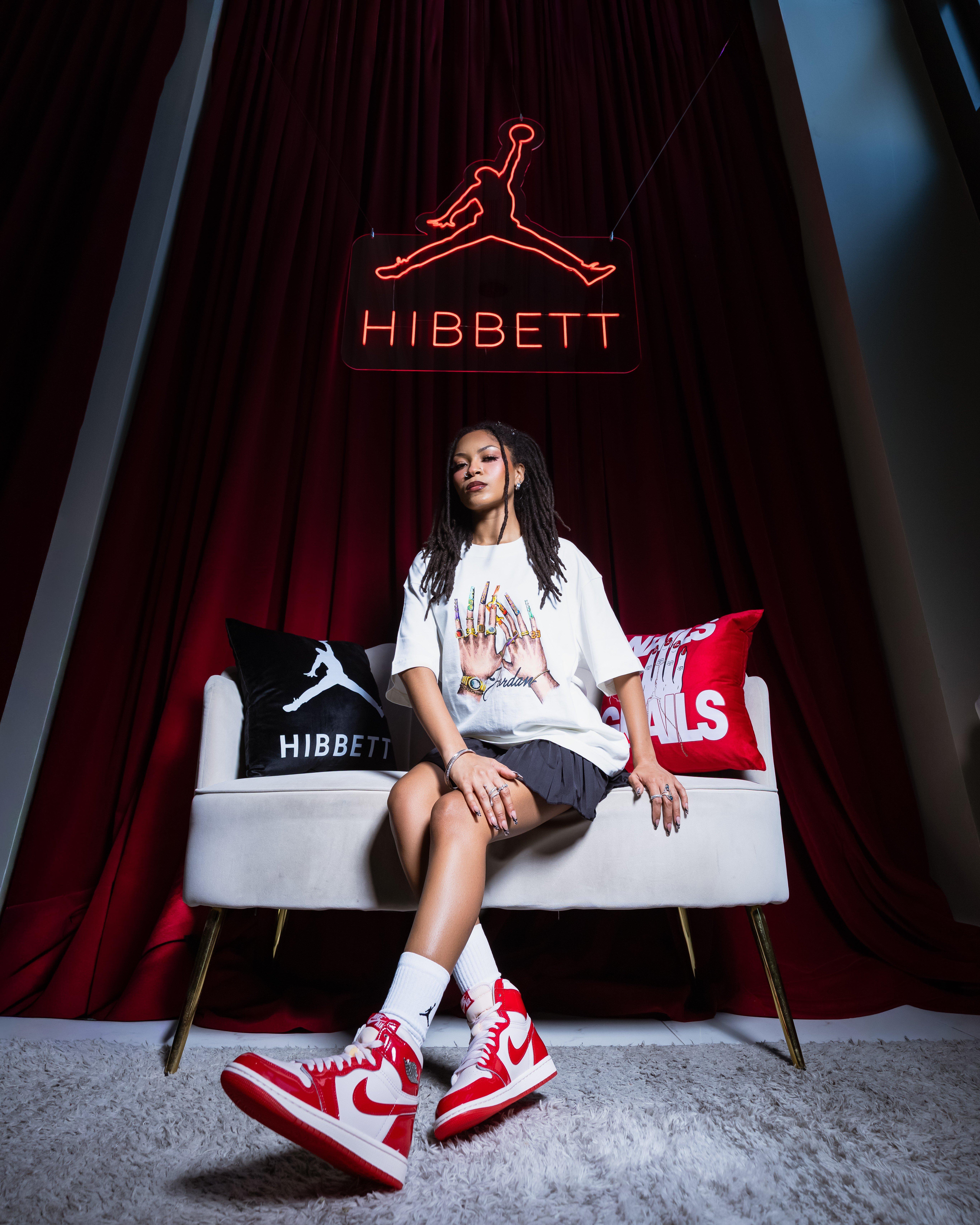Model seated beneath a red Hibbett Jumpman sign wearing a white Jordan graphic T-shirt, black pleated skirt and red-and-white Air Jordan 1 Retro High OG sneakers.