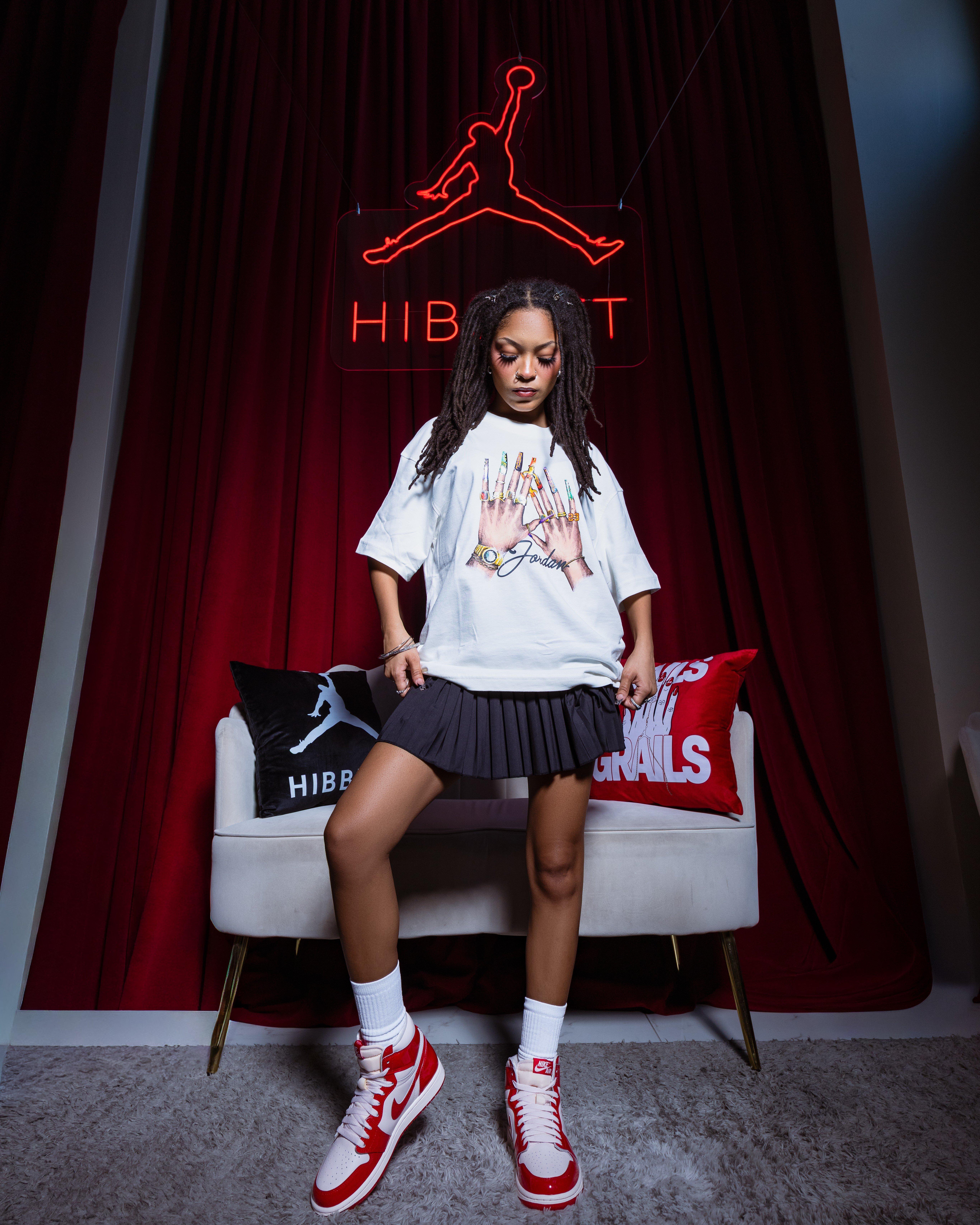 Model standing beneath a red Hibbett Jumpman sign wearing a white Jordan graphic T-shirt, black pleated skirt and red-and-white Air Jordan 1 Retro High OG sneakers.