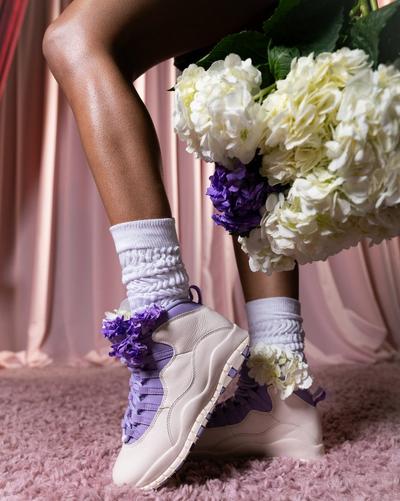 Close-up of Air Jordan 10 Retro Hydrangea sneakers in white and lavender with floral accents, styled with white scrunched socks and a bouquet of white and purple flowers.