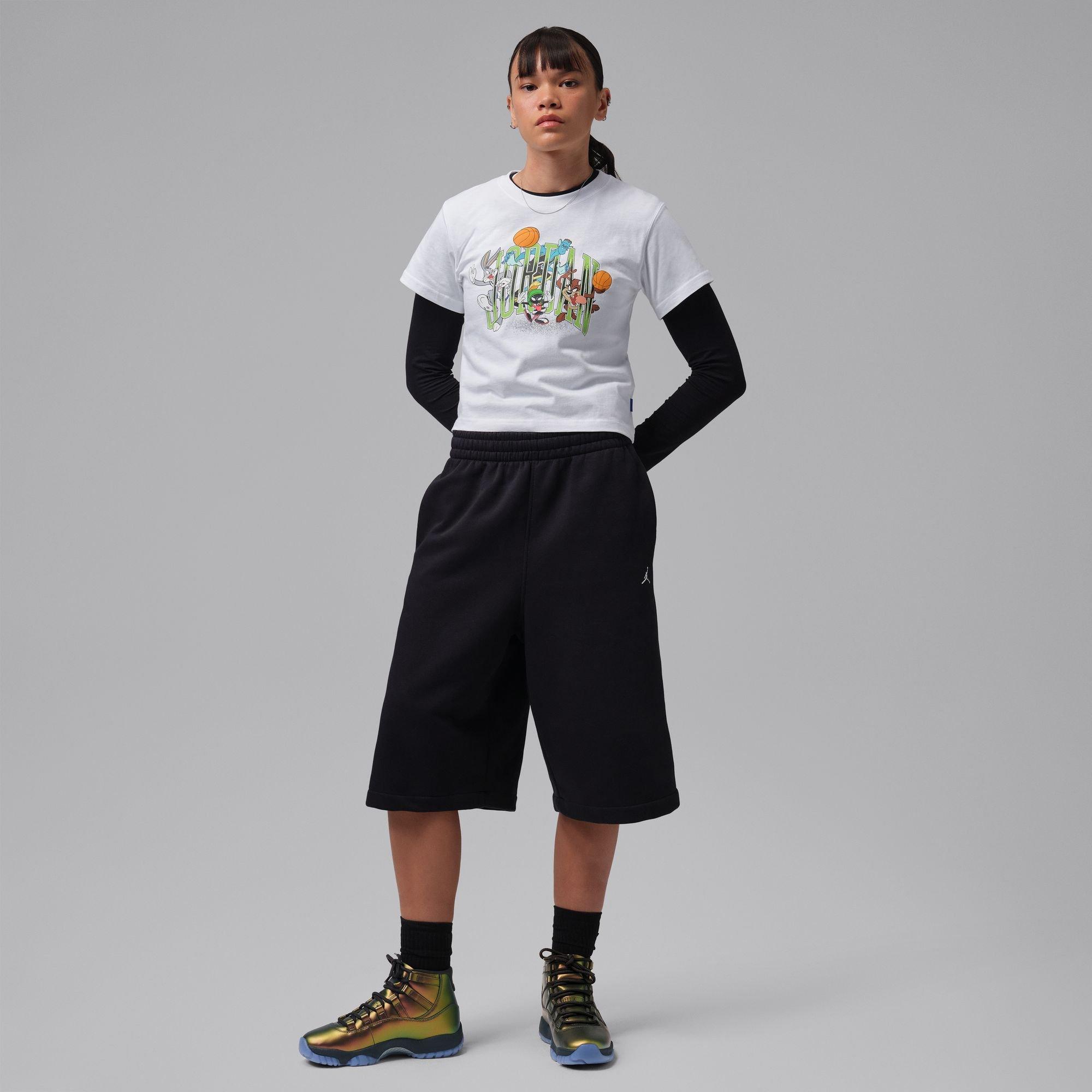 Person wearing a white Looney Tunes basketball graphic T-shirt, black wide-leg shorts and metallic green Jordan sneakers against a gray studio background.