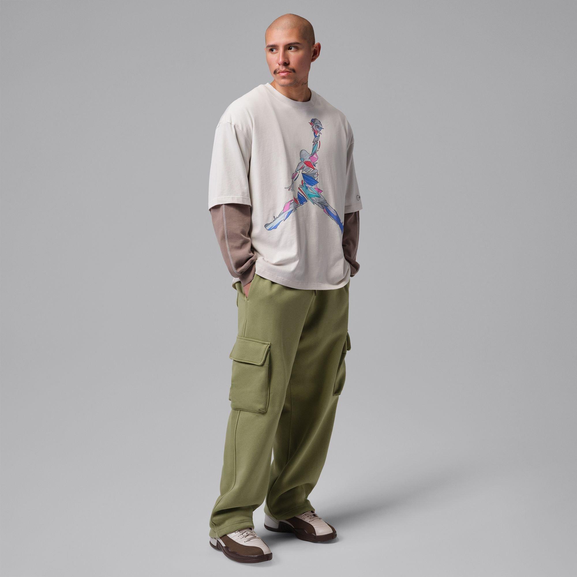 Model wears the Jordan 12 Retro &ldquo;Fireside&rdquo; with olive cargo pants and a layered graphic T-shirt against a gray studio backdrop.