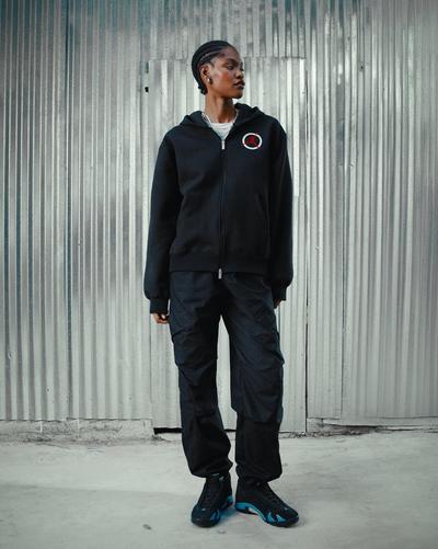 Person wearing a black Jordan tracksuit and Air Jordan 14 Retro sneakers in black with University Blue accents, standing in front of a corrugated metal wall.