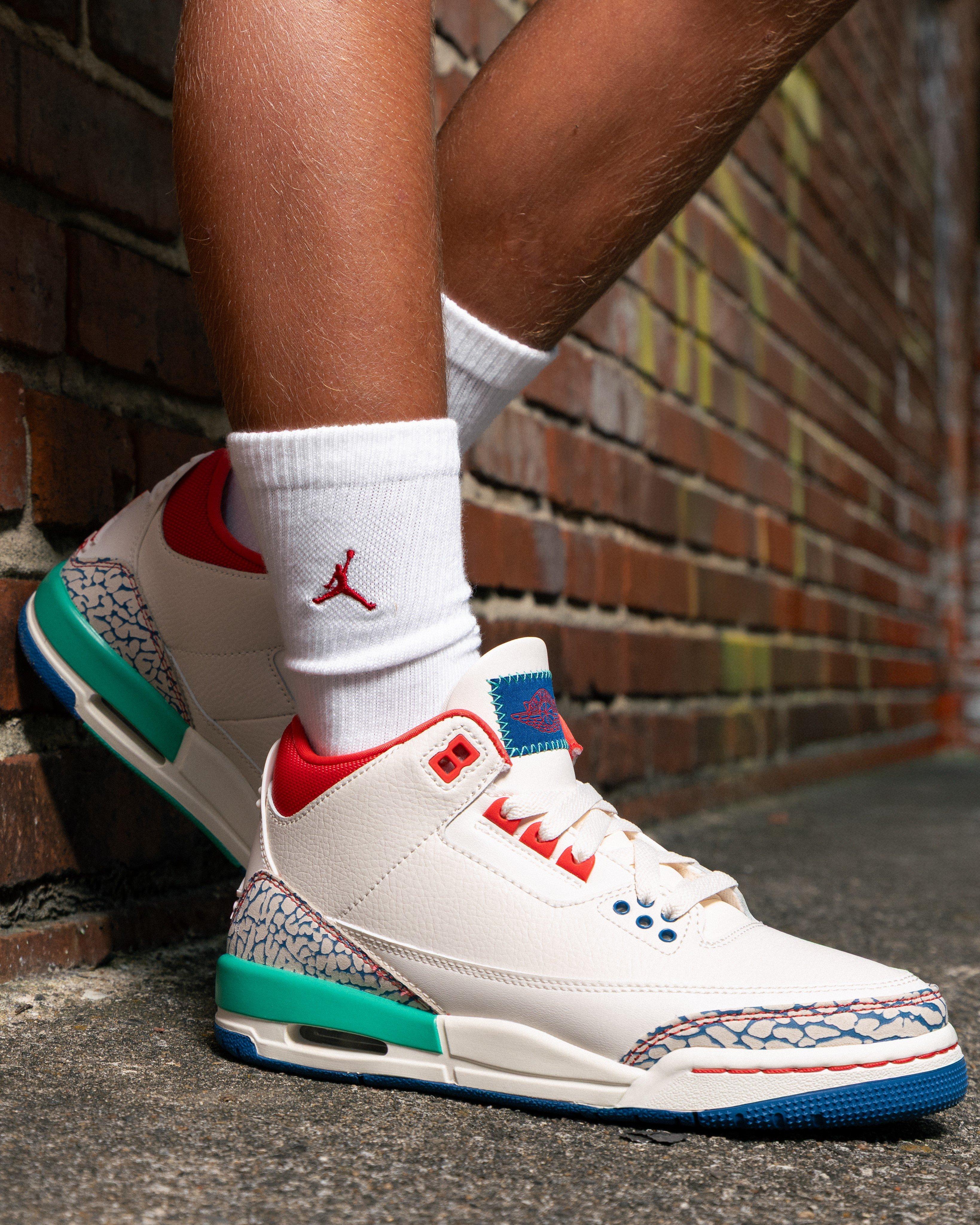 Close-up of Jordan 3 Retro Chalk and True Blue kids sneakers with white leather, red accents, teal heel details, blue outsole and elephant print overlays, worn with white Jordan crew socks against a brick wall.