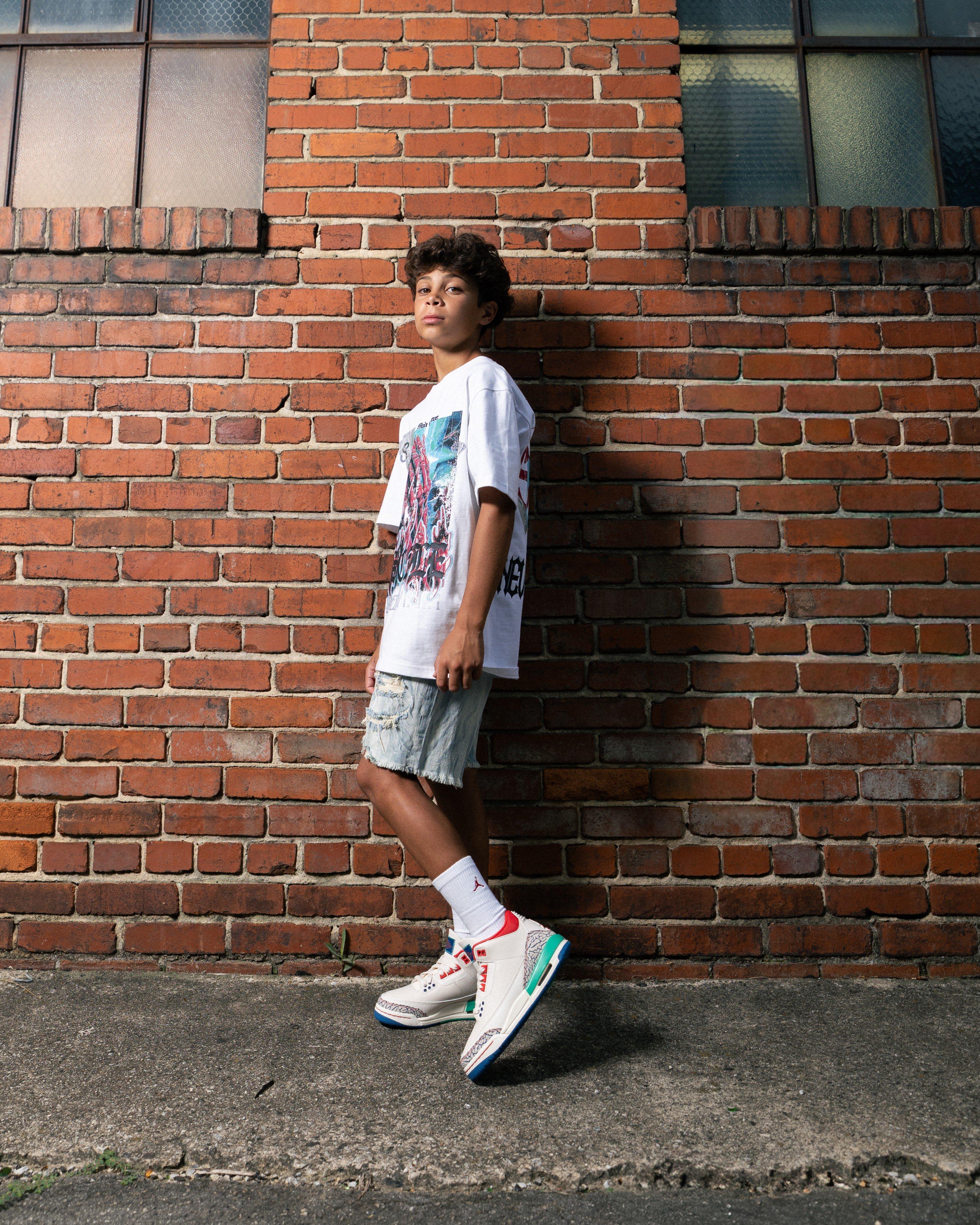 Person standing in front of a brick wall wearing Jordan 3 Retro Chalk and True Blue kids sneakers, white Jordan socks, light denim shorts and an oversized white graphic T-shirt.