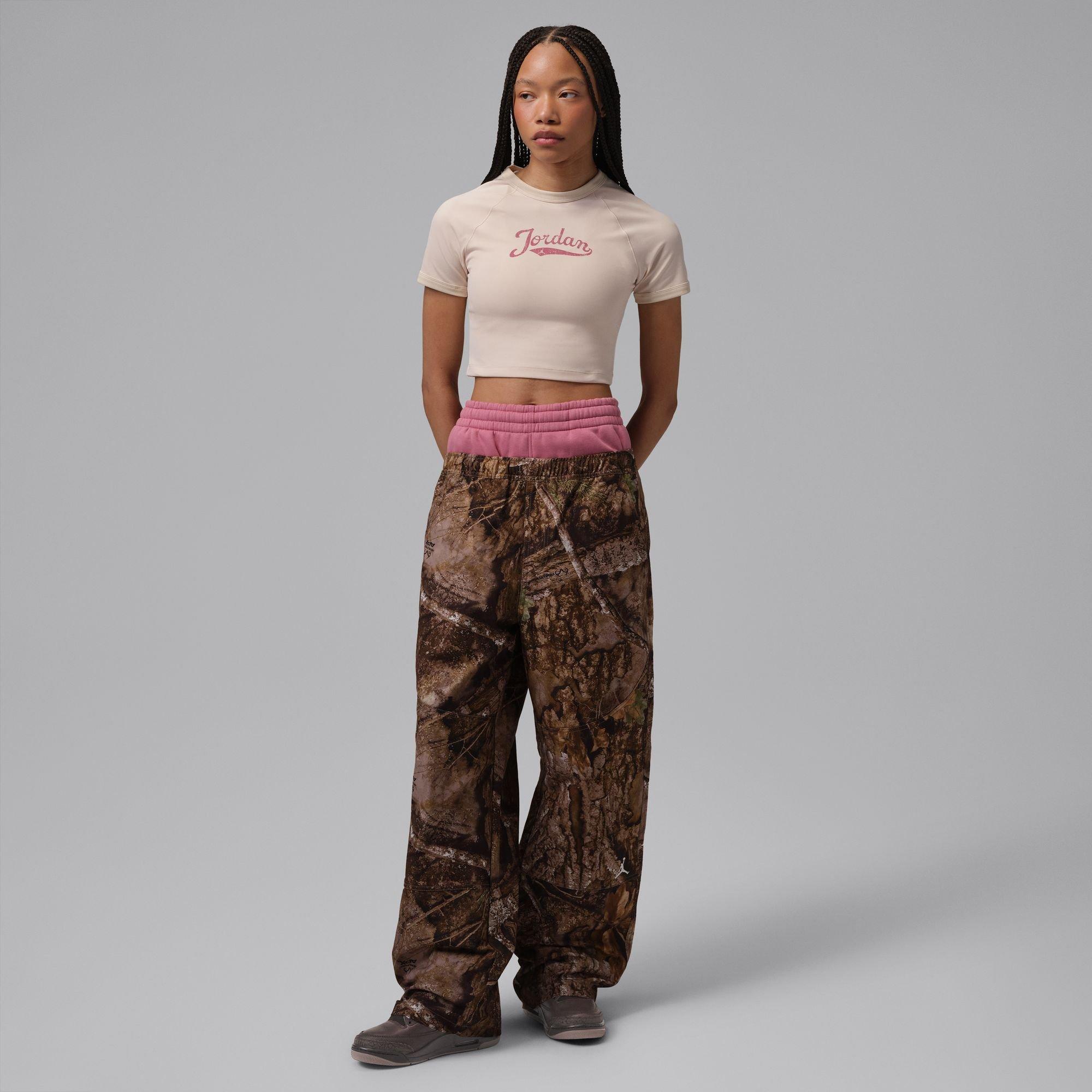 Model wearing a beige Jordan crop top, layered pink and camouflage pants and brown Air Jordan sneakers against a gray studio background.
