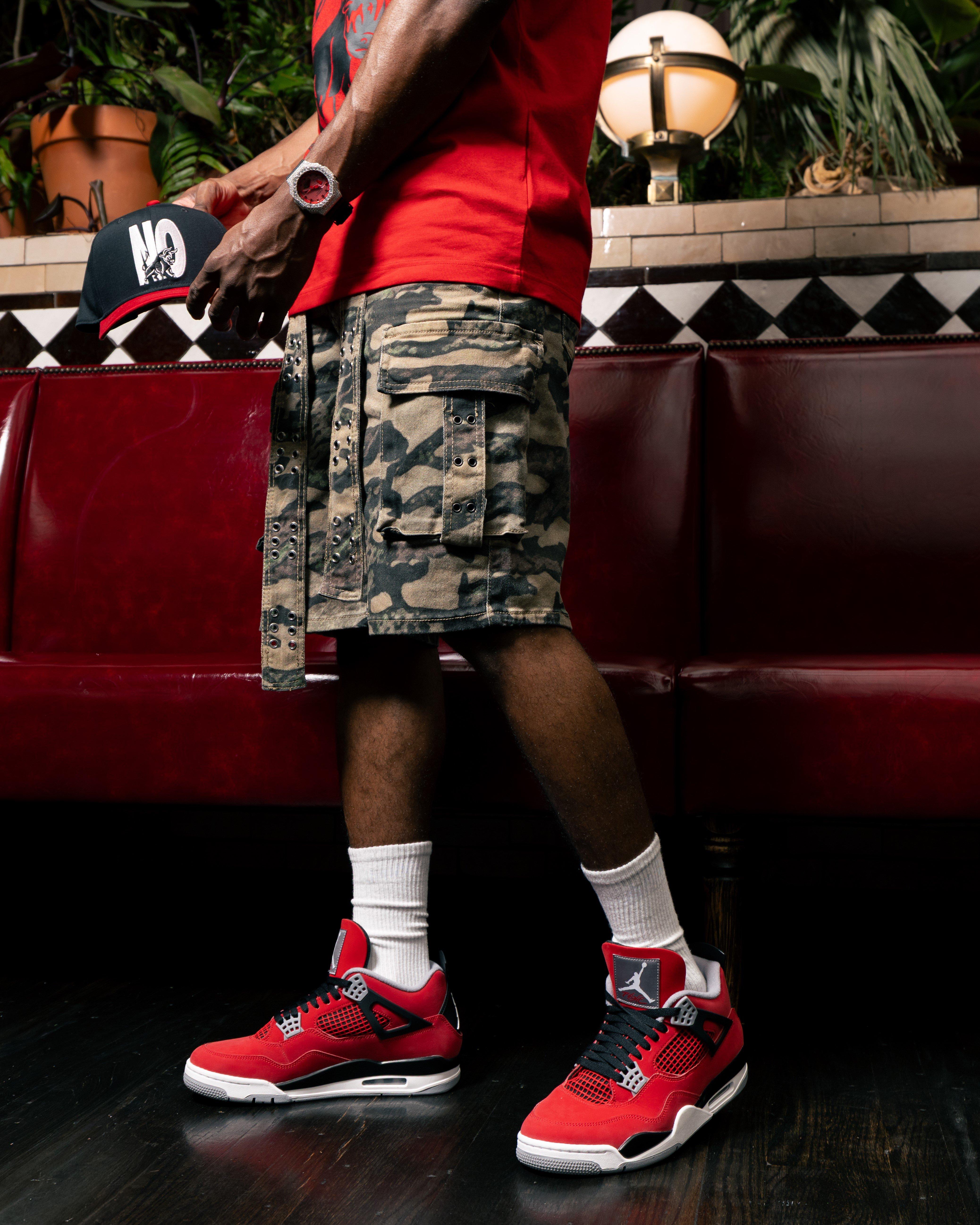 Person in camo shorts holding a black and red cap, wearing red sneakers and watch.