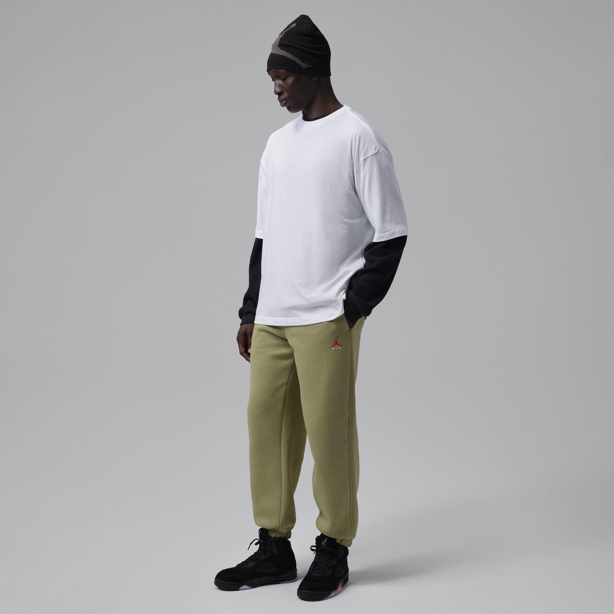 Model wearing a white layered T-shirt, olive Jordan sweatpants, black beanie and black Air Jordan 5 Retro Halloween sneakers against a gray studio background.