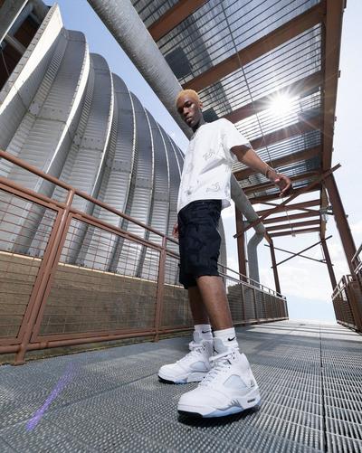 Person standing on an industrial metal walkway wearing Air Jordan 5 Retro White Metallic sneakers with an all-white upper, reflective tongue and icy translucent outsole.
