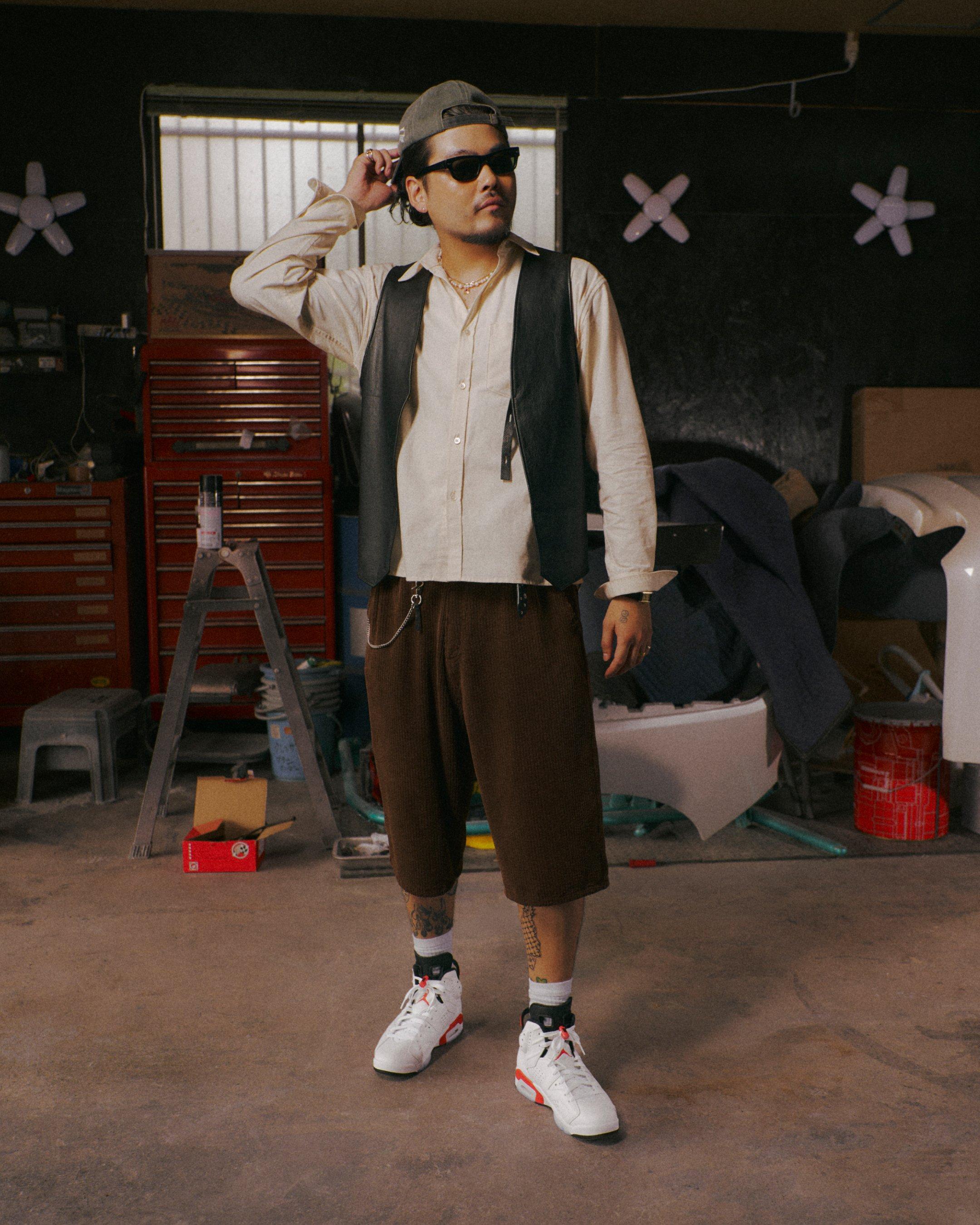 Person wearing a cream shirt, black vest, brown cropped pants and Jordan 6 Retro White Infrared sneakers in a workshop.