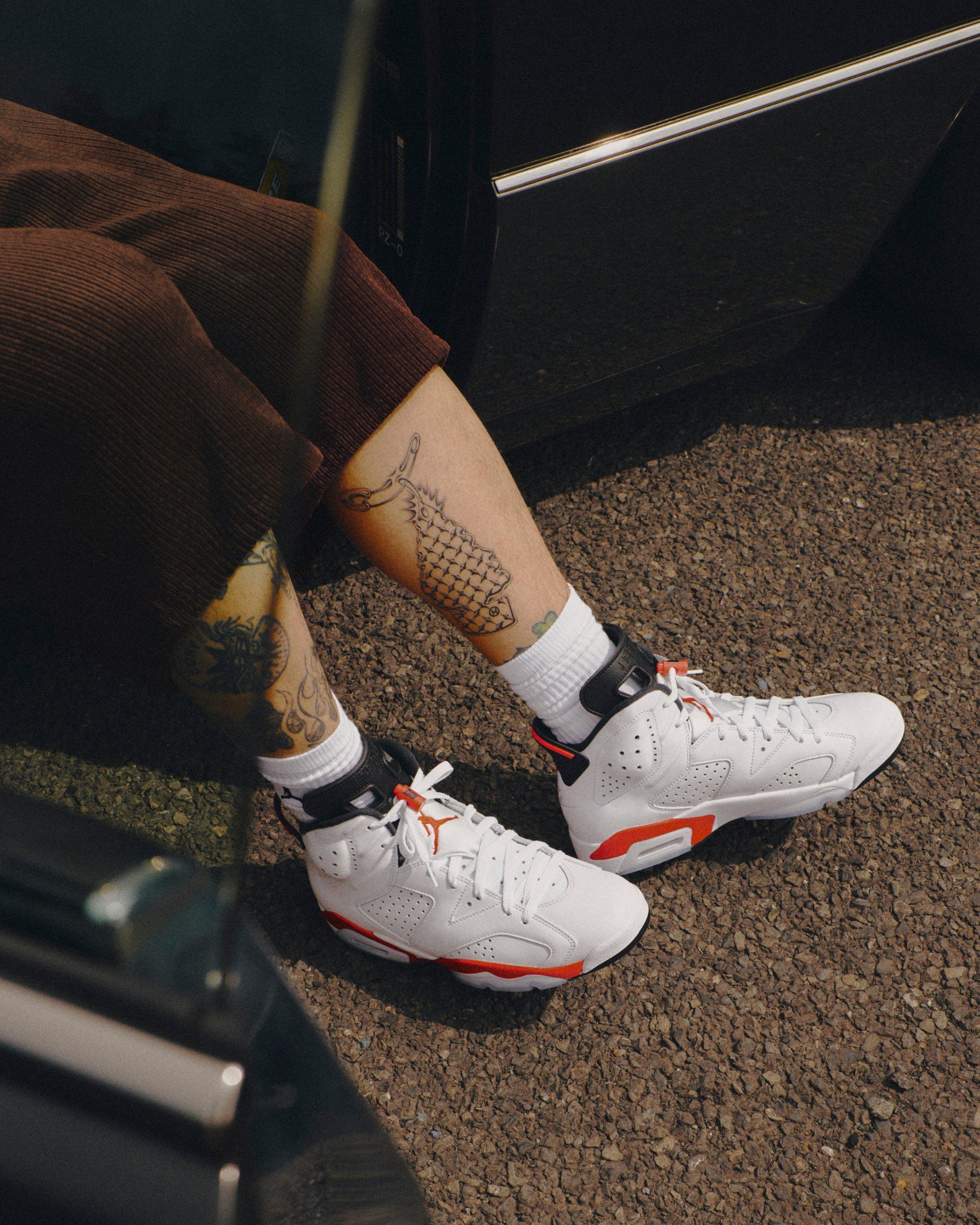 Jordan 6 Retro White Infrared sneakers worn with white crew socks and brown shorts while seated on a textured floor.