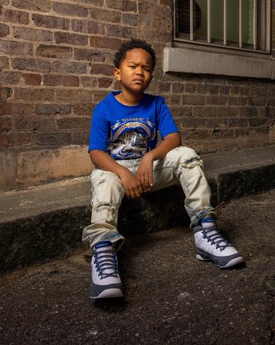 Child sitting on a city sidewalk wearing Air Jordan 9 Retro Flint Grey sneakers featuring a white upper, grey mudguard and blue details, paired with light-wash jeans.