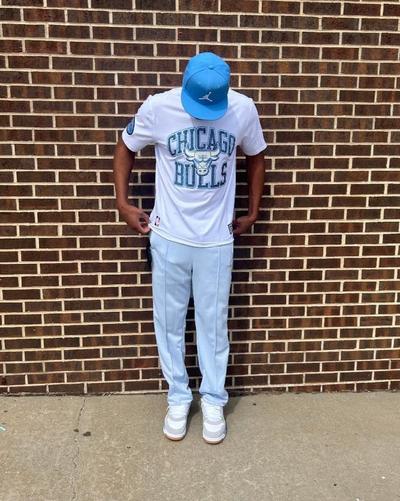 A full-body shot of a person standing in front of a brick wall, wearing a light blue baseball cap, a white t-shirt with "CHICAGO BULLS" in light blue lettering, light blue track pants, and light gray and white sneakers. The person's head is tilted slightly down, and their hands are holding the waistband of their pants.
