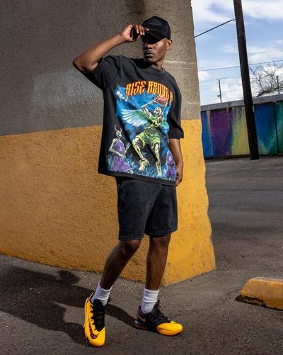 Model wearing a black Rise Above It basketball graphic T-shirt, black shorts and mismatched yellow-and-black Nike basketball shoes outdoors.