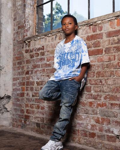 Kid wearing a blue graphic T-shirt, distressed jeans and white sneakers, leaning against a brick wall.