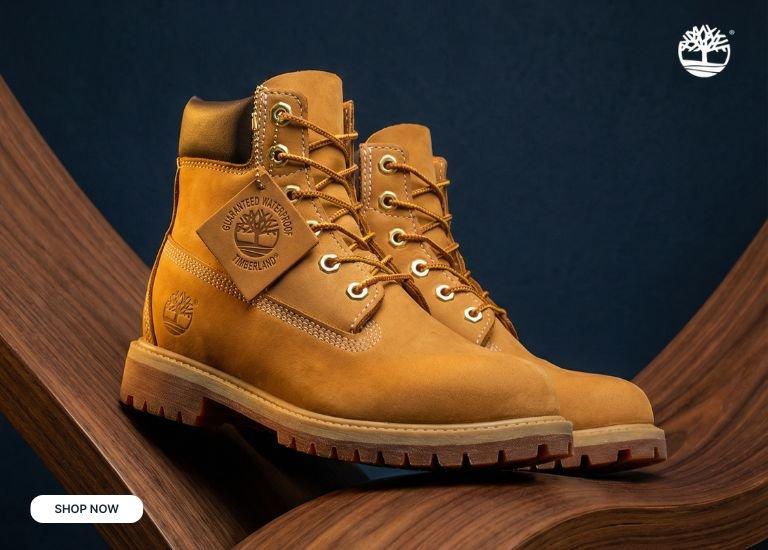 An advertisement for classic Timberland 6-Inch Premium Waterproof Boots in the iconic Wheat color. The image shows a pair of boots resting on a curved, dark wood surface against a textured navy blue background. The boots feature gold eyelets, two-tone laces, a dark brown padded collar, and the Timberland tree logo embossed on the side. A SHOP NOW button is in the top left corner, and the white Timberland logo is in the top right.