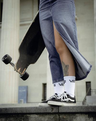 Close-up of a person holding a worn skateboard while wearing black Vans sneakers, white Vans crew socks and a denim skirt with a side slit.