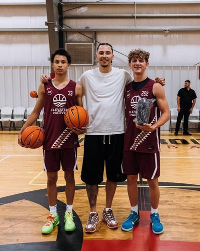 Two basketball players in maroon Elevated Native jerseys stand with basketballs beside a person in a white T-shirt on an indoor court, with one player holding a wrapped pair of shoes.