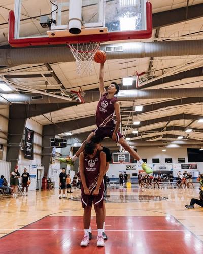 Basketball player in a maroon Elevated Native jersey leaps over a teammate to attempt a dunk during an indoor basketball event, with spectators and players watching from the sidelines.