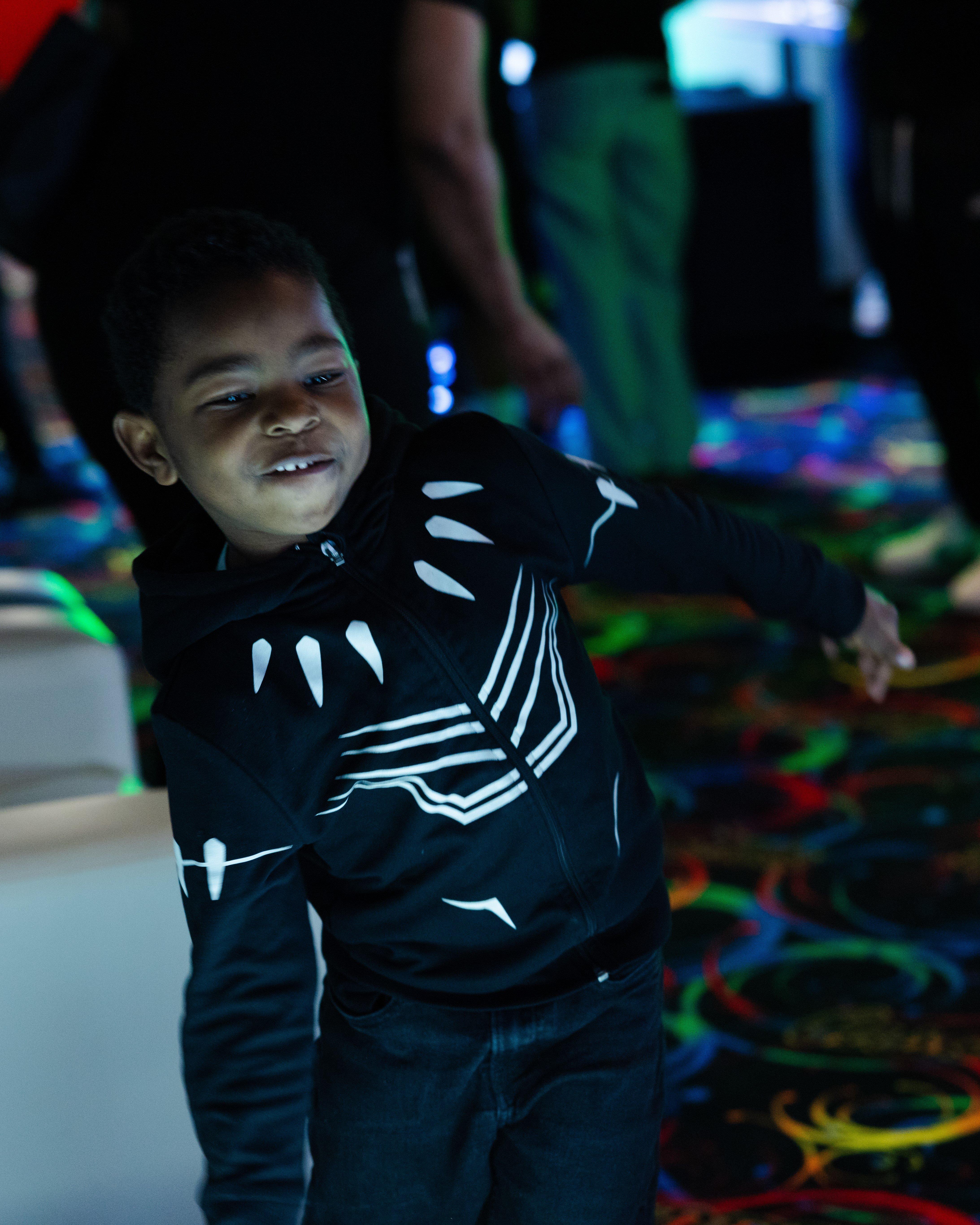 A candid shot of a young boy in a black "Black Panther" themed hoodie dancing on a colorful, patterned carpet under low, atmospheric event lighting.