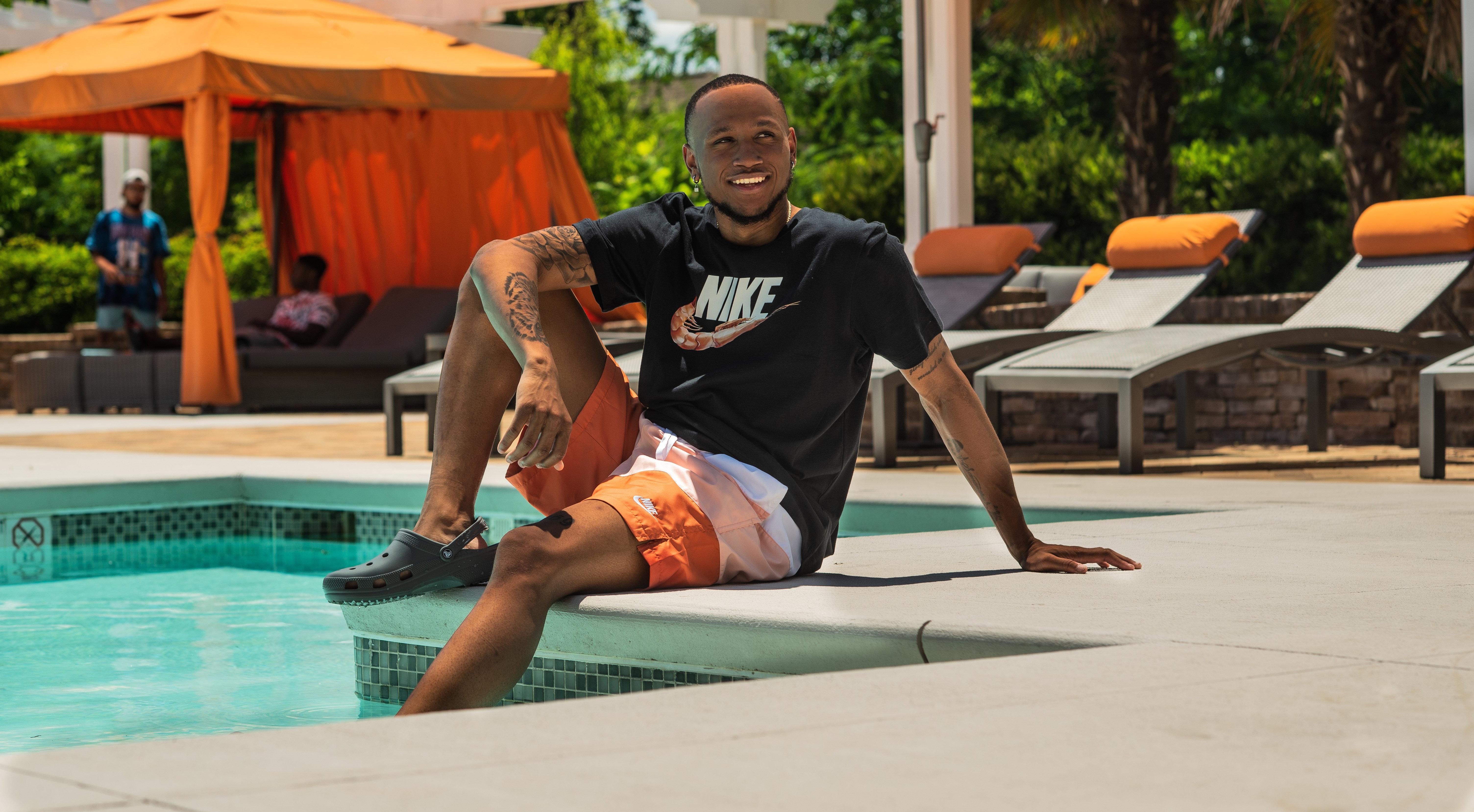 Man by pool in Nike Shrimp Swoosh tee, orange shorts and black Crocs clogs