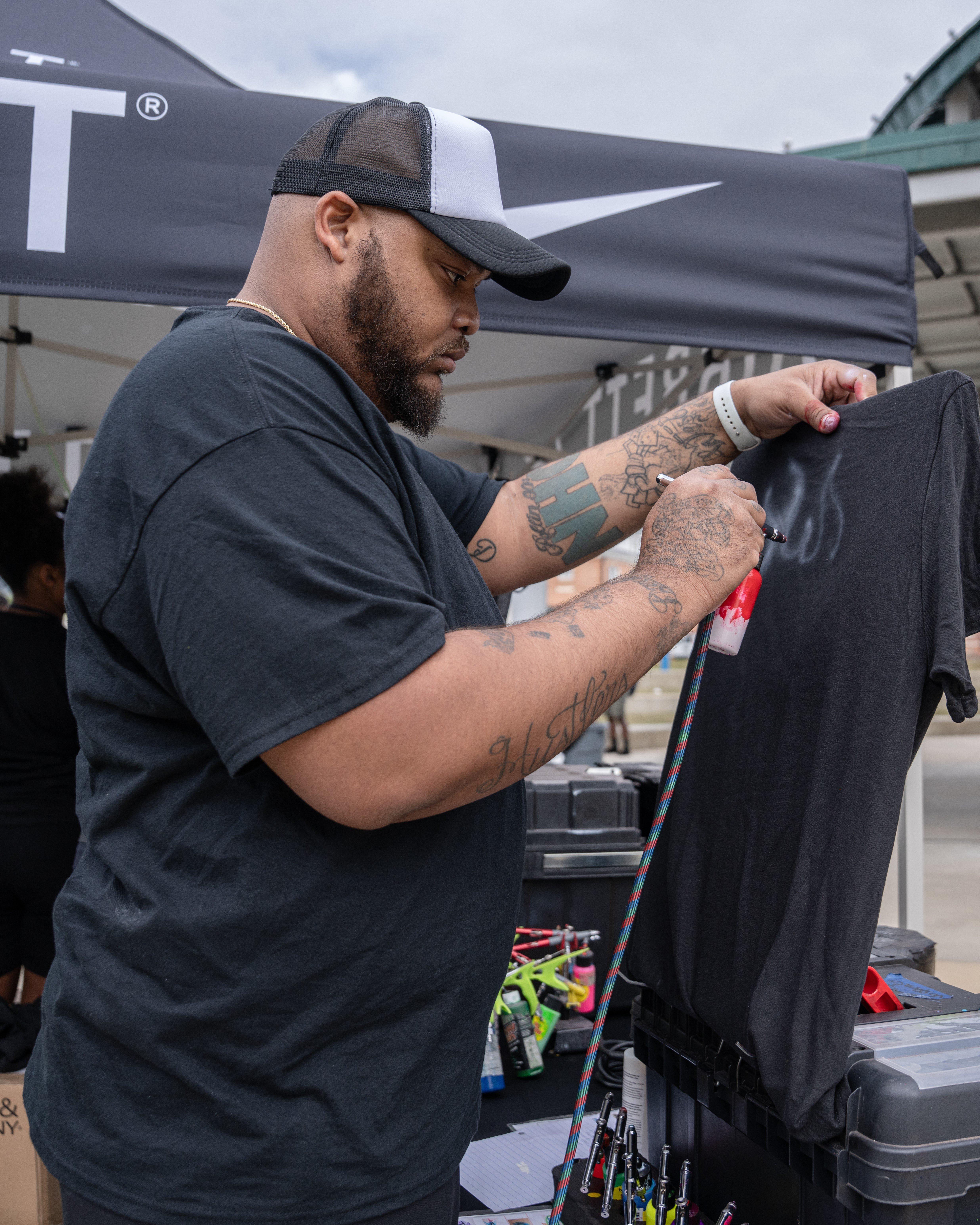 A man with a beard, tattoos, and a two-tone baseball cap is airbrushing a design onto a black t-shirt.