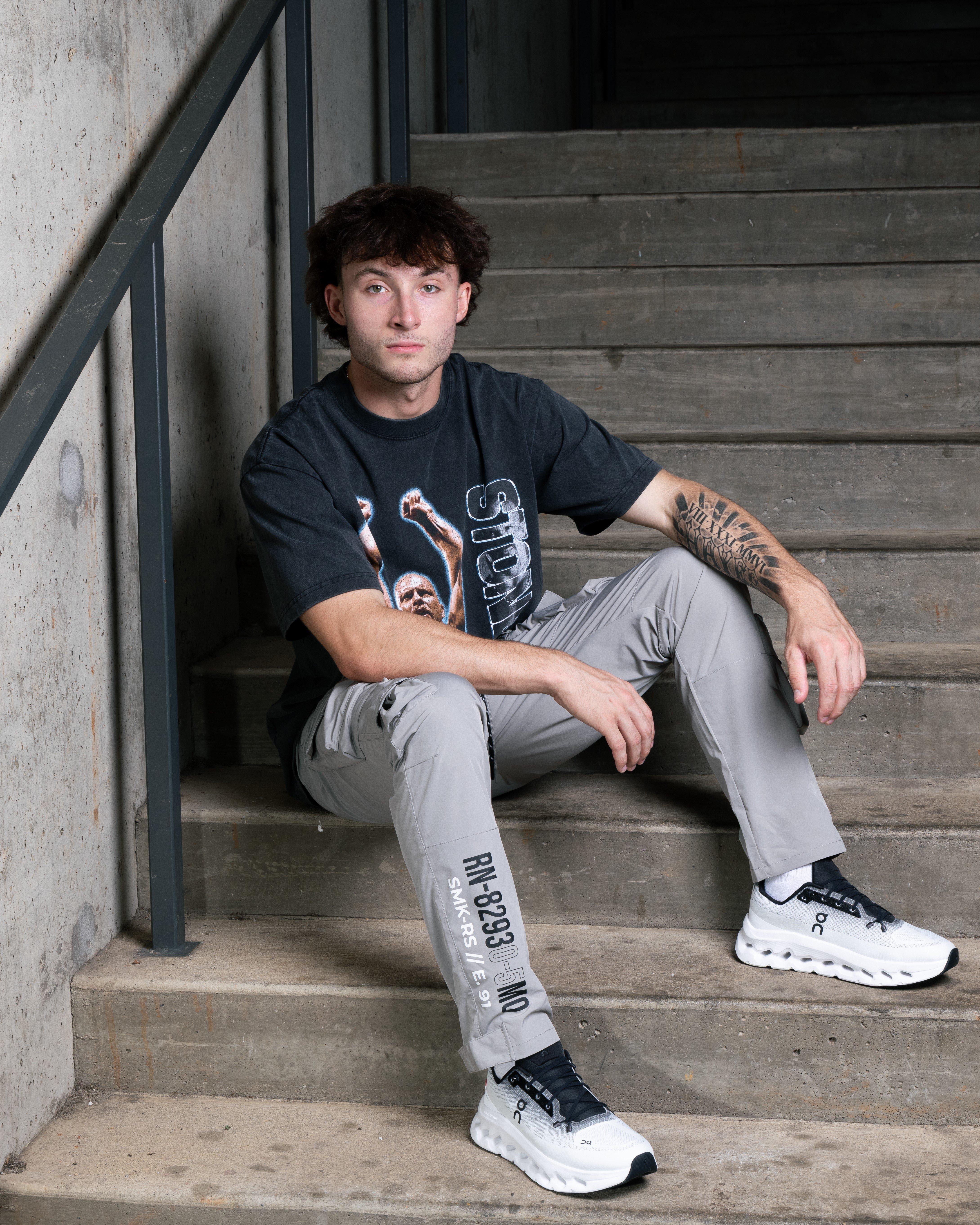 A full-body shot of a man with curly brown hair sitting on concrete stairs. He is wearing a dark gray graphic t-shirt, light gray pants with black text on the leg, and white and black On sneakers. He has a tattoo on his left forearm.