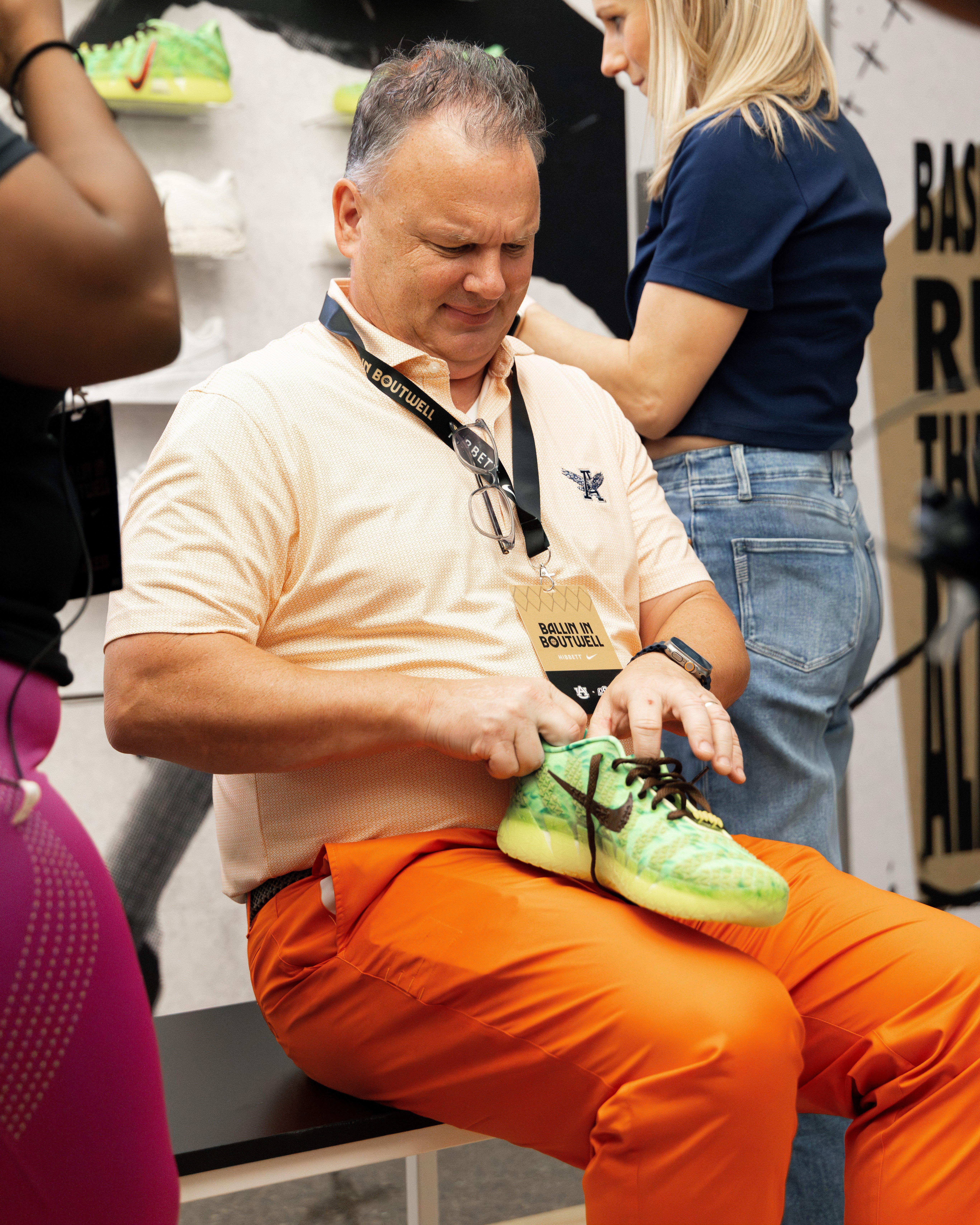 A group photo of the HuffmanaA man wearing a peach polo and bright orange pants is sitting down, looking at a pair of neon green and black Nike shoes in his hands. He has a lanyard that reads BALLIN IN BOUTWELL. High School Girls Basketball team and their coaches, standing on the court in matching green tracksuits while holding basketballs.