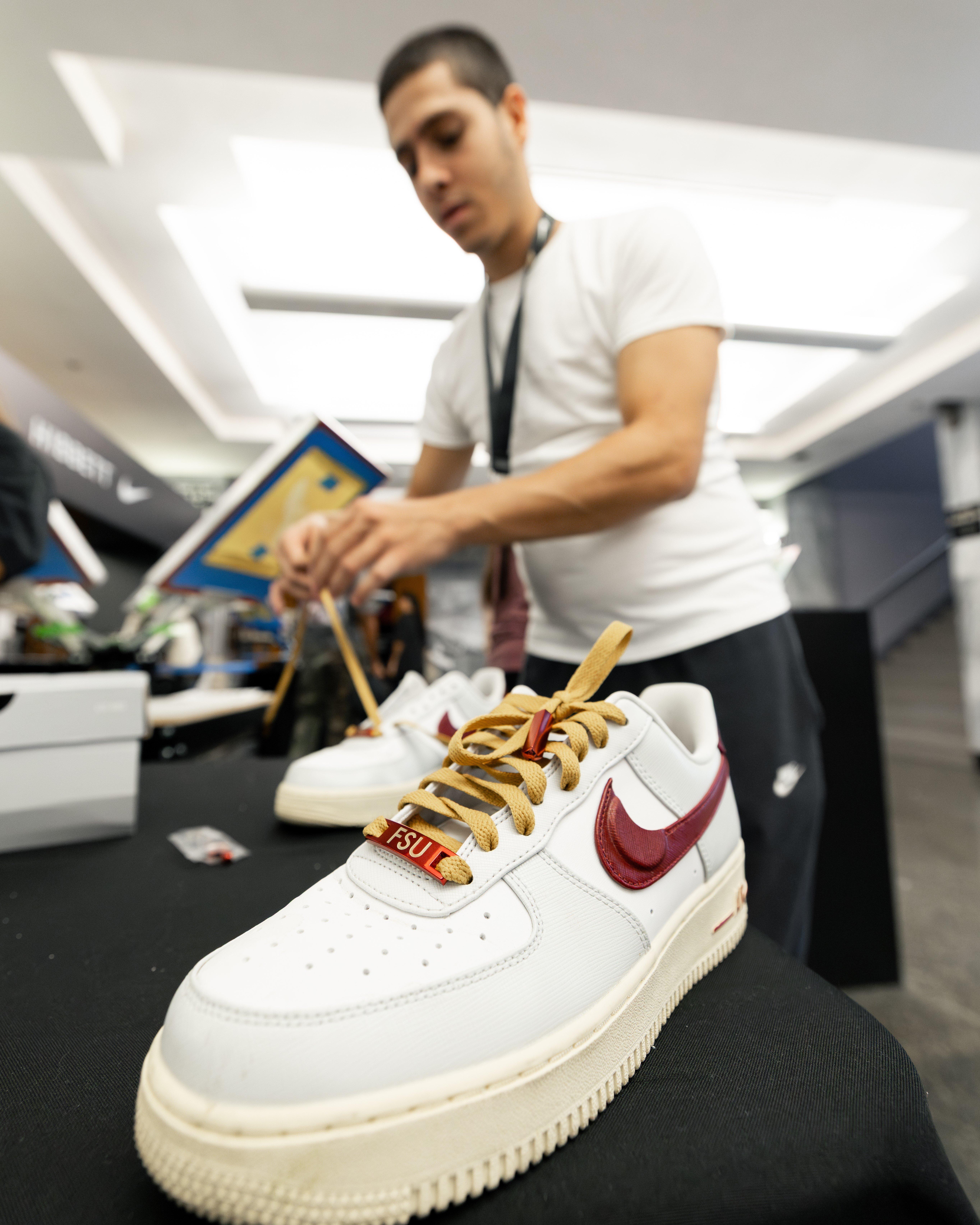 A close-up of a white Nike Air Force 1 sneaker with a maroon swoosh and gold laces, displayed on a table. In the blurry background, a man is working on another shoe. The shoe has an FSU tag near the toe box.
