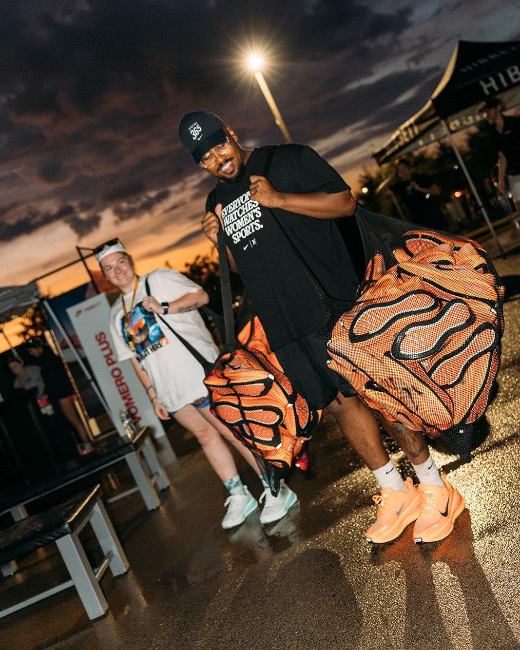 A man wearing a black t-shirt and a baseball cap smiles at the camera while holding two large orange mesh bags with a wavy pattern on them. Another person is visible behind him. The ground is wet, suggesting it might have rained. The scene is lit by a streetlamp at dusk.