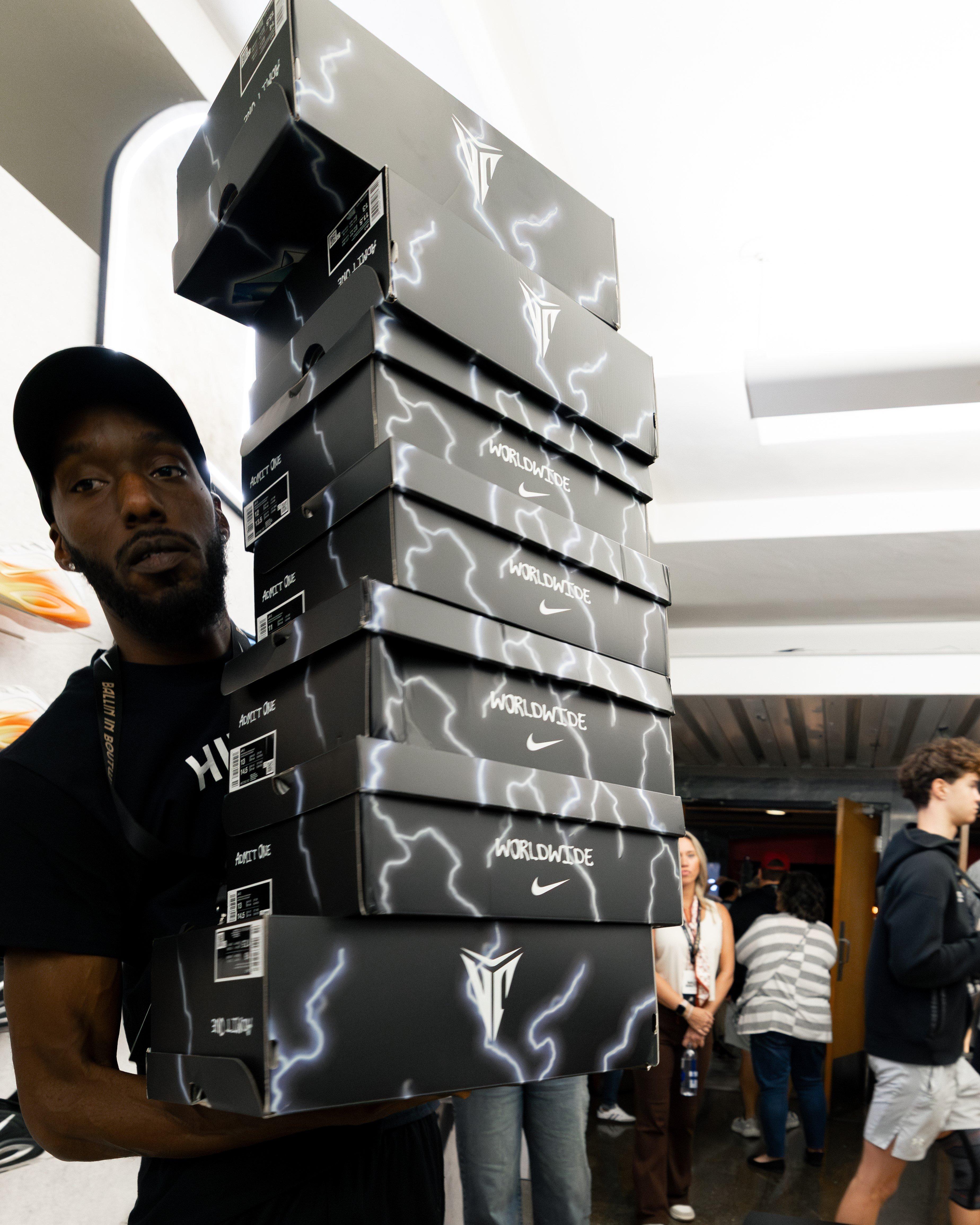A tall man wearing a black cap is holding a large, stacked pile of black shoe boxes decorated with a white lightning bolt design. The boxes have WORLDWIDE and HIBBETT branding.
