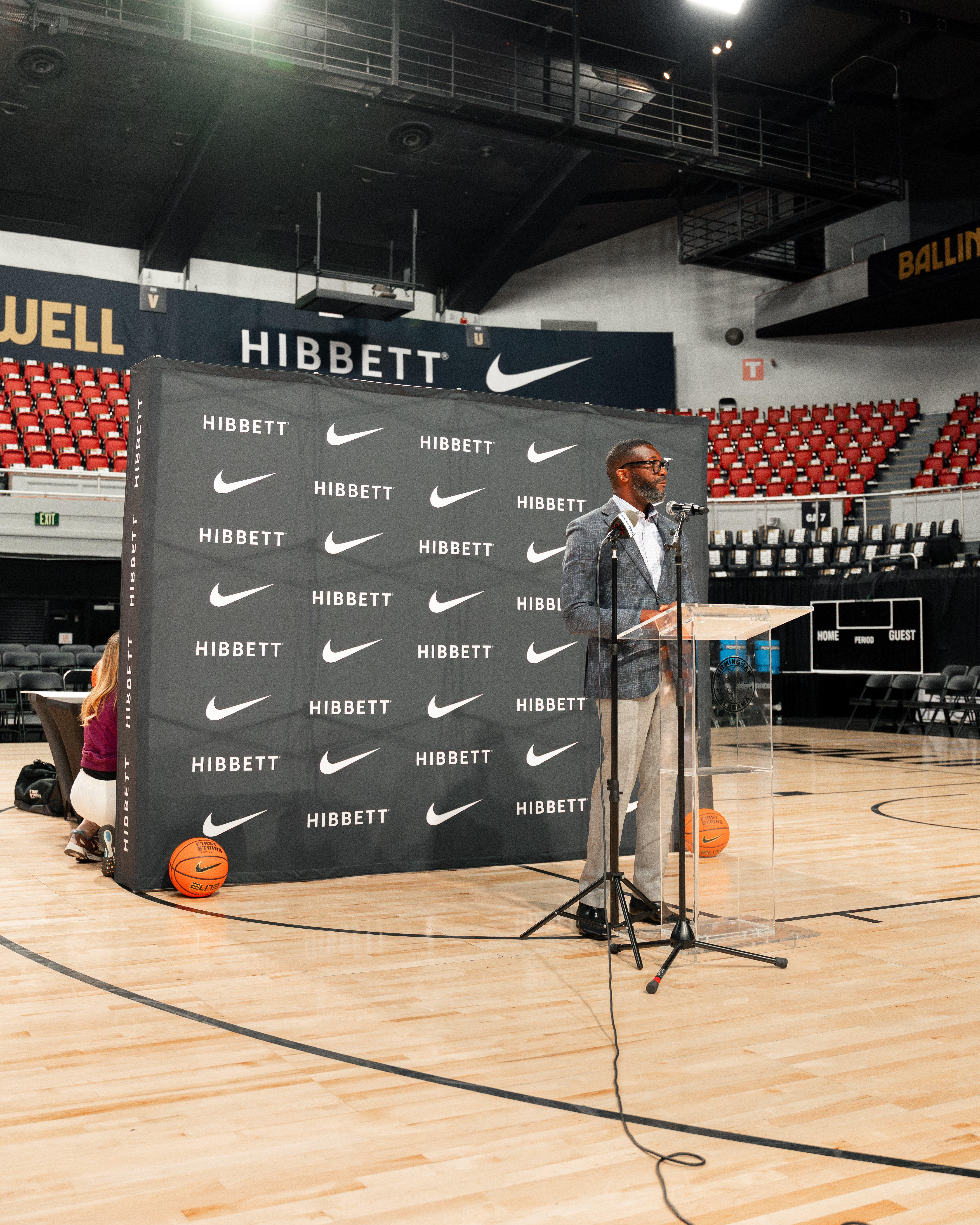 A man in a suit jacket is speaking at a podium on a basketball court in a large arena. Behind him is a tall black backdrop featuring the HIBBETT logo and Nike swoosh repeated across it.