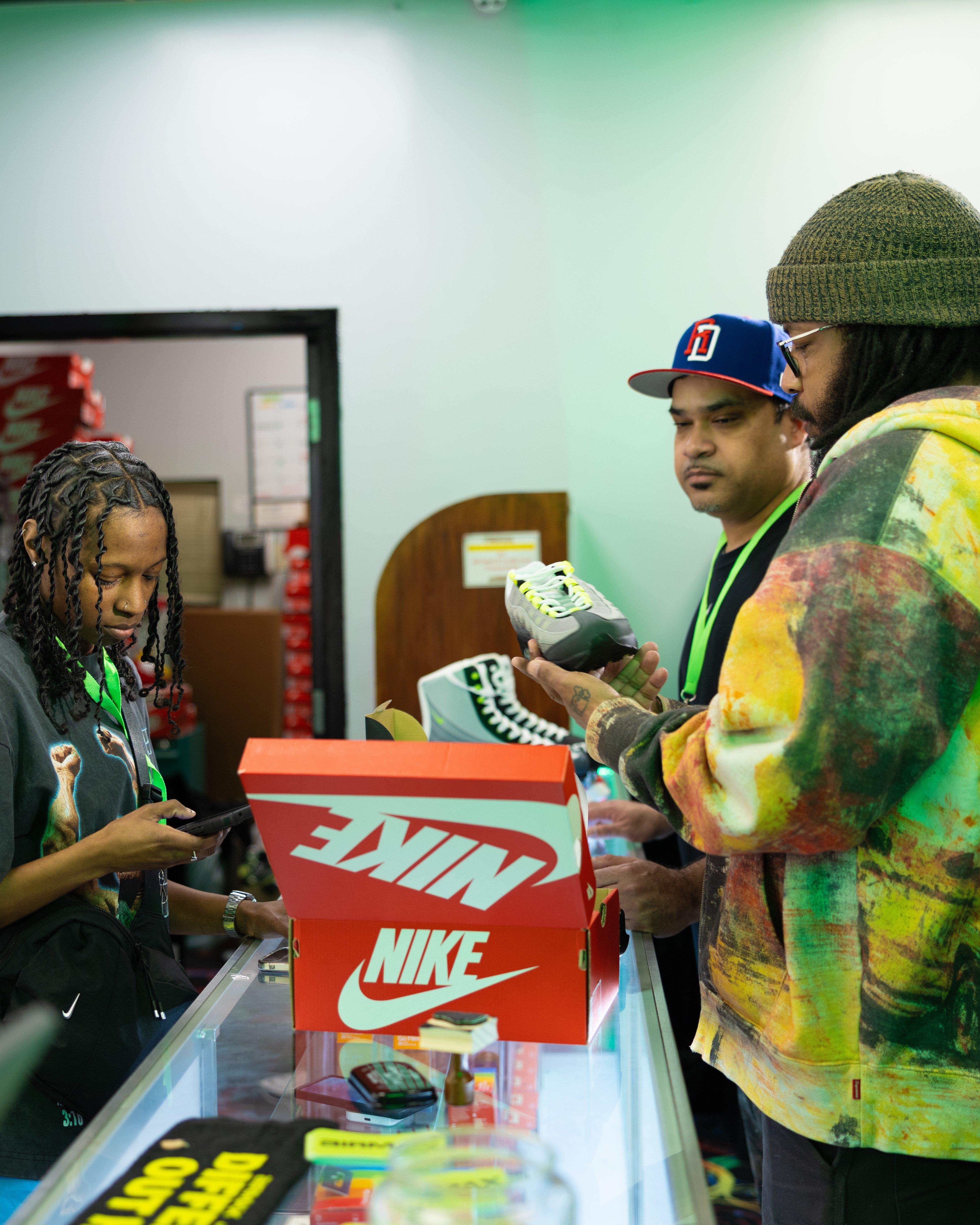 A retail setting where a man in a colorful hoodie and beanie examines a Nike Air Max 95 "Neon" sneaker. Beside him, a man in a blue baseball cap looks on, and a store employee in a grey shirt is focused on a smartphone.