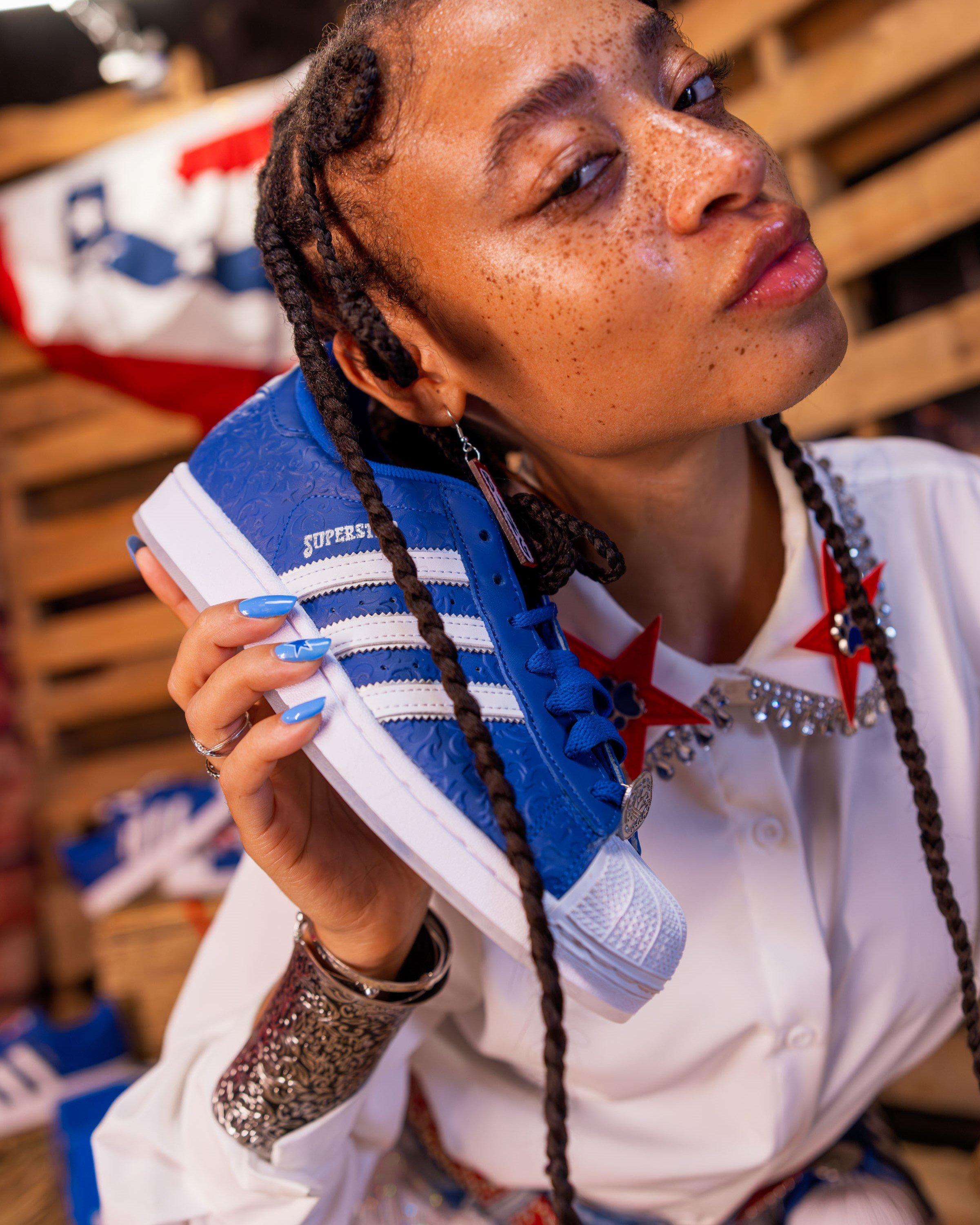 A close-up shot of a model with freckles and braided hair. She is holding a blue and white Adidas Superstar shoe up to her ear like a phone. She is wearing a silver cuff bracelet and a rhinestone necklace with a red star pendant.