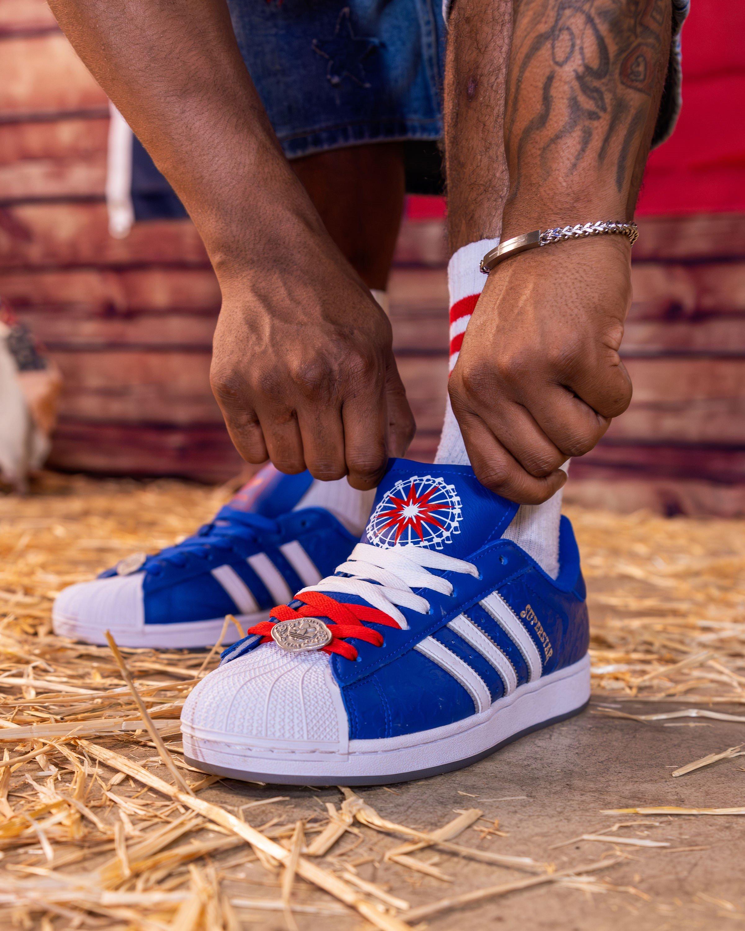 A person is tying the red and white laces on a blue Adidas Superstar shoe. The shoe has a round, white and red logo on the tongue and a silver coin charm on the laces. The person is wearing a silver bracelet and white socks with red stripes. The background is a bed of hay.