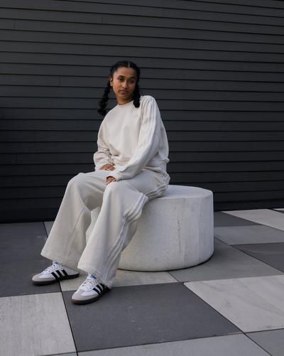 Model seated on a white bench wearing a cream adidas jogger set and white adidas Samba sneakers with black stripes against a dark paneled wall.