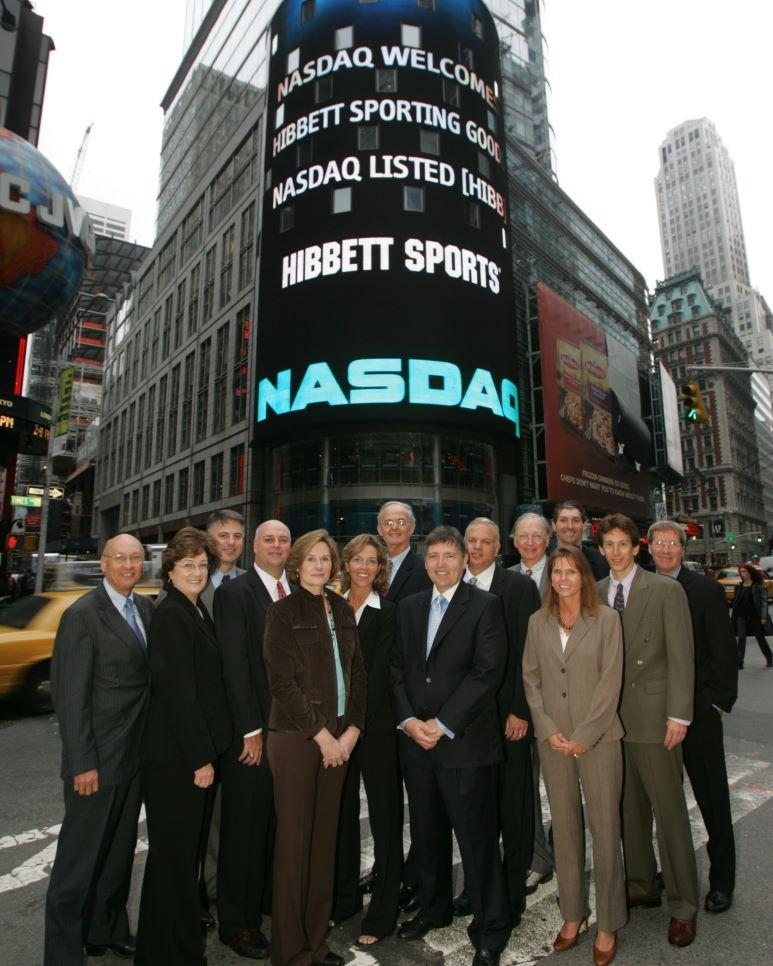  A group of people on a city street in front of the NASDAQ MarketSite building in Times Square, New York City. The large, curved LED display on the building prominently features the NASDAQ logo and the text "HIBBETT SPORTS."