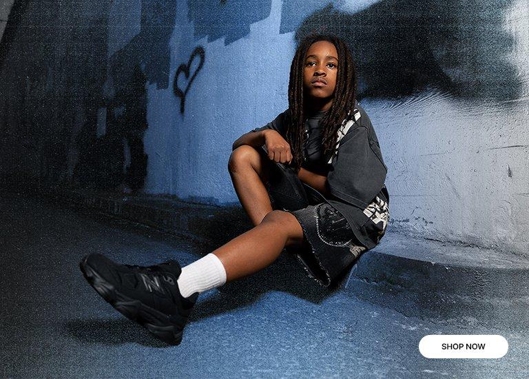 Lifestyle banner featuring a model seated against a blue-toned graffiti wall with a heart illustration. He is wearing a dark charcoal graphic tee, matching patterned shorts, white crew socks, and triple black New Balance 9060 sneakers. A white Shop Now button is located in the bottom right corner.