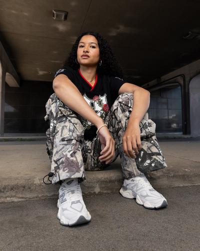 Young woman modeling Smoke Rise Roses Oversized Jersey - Black, Smoke Rise Wide Leg Woven Pants - Camo, and New Balance 9060 Sea Salt