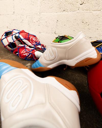 Close-up of Nike Air Zoom 90 sneakers in white and blue with gum soles, placed on pavement next to a net of colorful soccer balls.