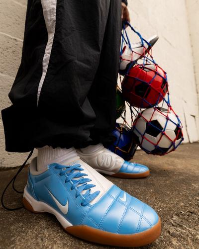 Person wearing Nike Air Zoom 90 sneakers in blue and white with gum soles, holding a net filled with soccer balls against a concrete wall
