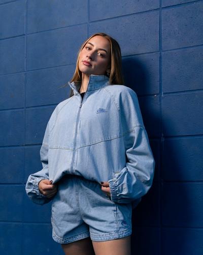 Model wearing a light-wash Nike denim zip jacket and matching shorts while standing against a blue brick wall.