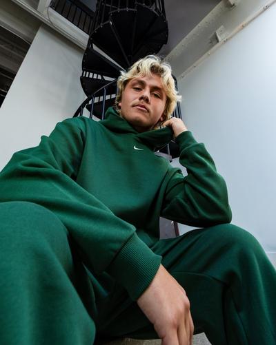 Model seated near a black spiral staircase wearing a matching dark green Nike hoodie and sweatpants.